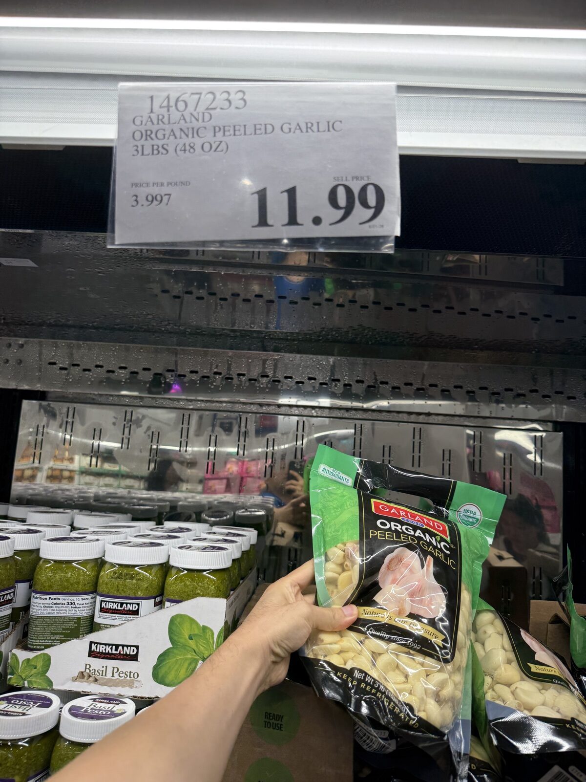A bag of Garland organic peeled garlic held up in front of the refrigerated case at the Leesburg, Virginia Costco, with the shelf sign showing 3 lbs for $11.99