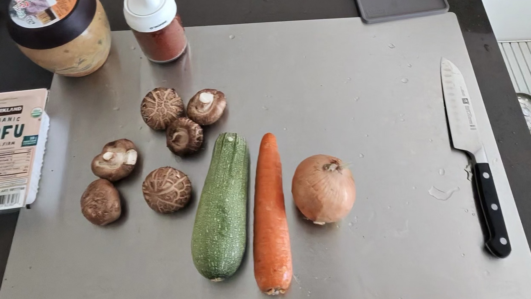 Whole shiitake mushrooms, a zucchini, a carrot and an onion lined up on a cutting board beside a package of tofu and a jar of doenjang