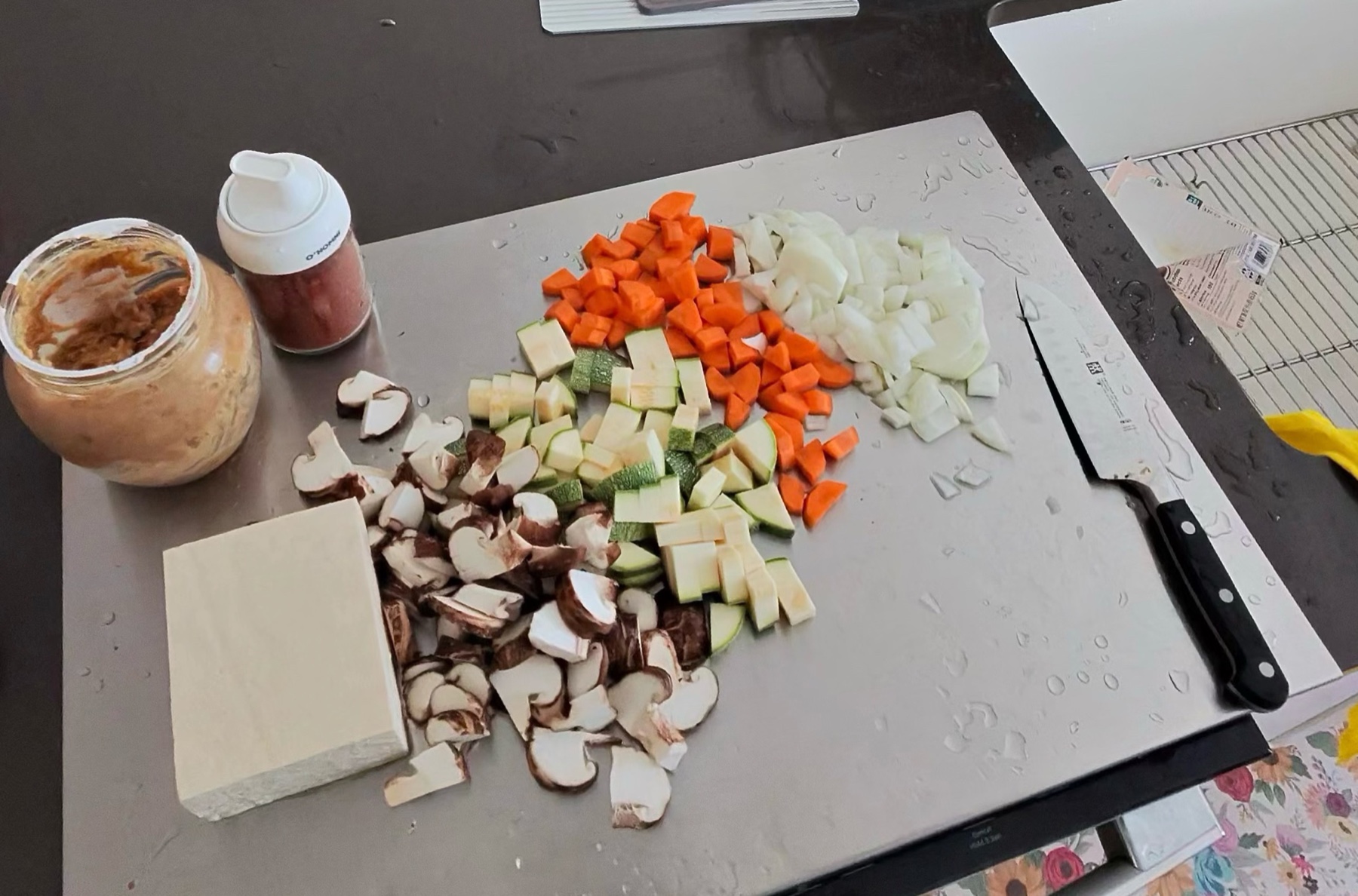 Diced shiitake mushrooms, zucchini, carrot and onion on a cutting board with a block of tofu and an open jar of doenjang