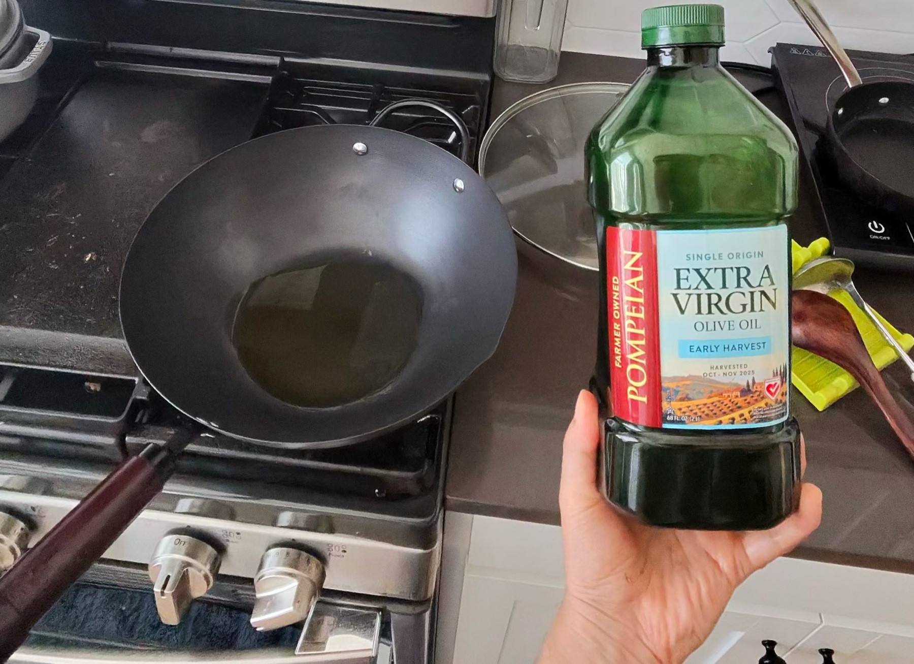 Olive oil being poured into a carbon steel wok on a gas stove, with the bottle held beside it
