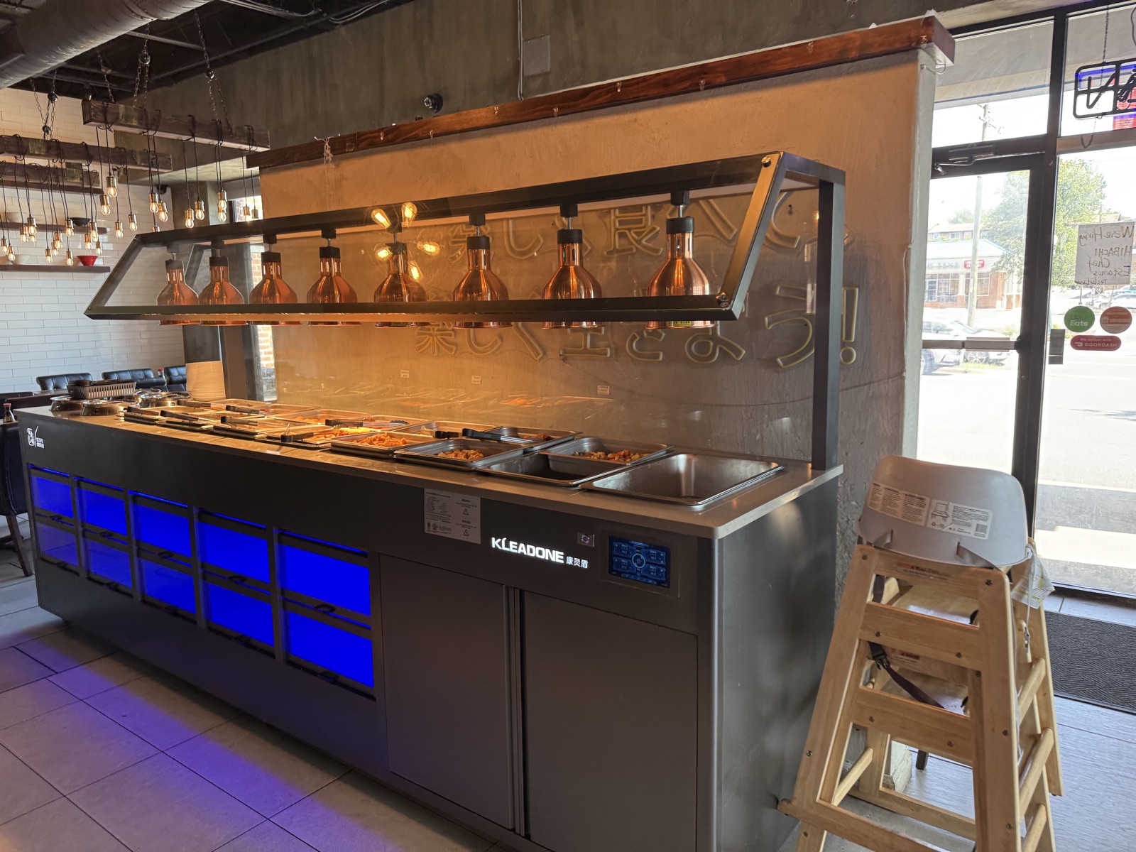 The full hot food bar setup at Sushi Oishii, a long steel counter with warming lamps and a row of steam trays