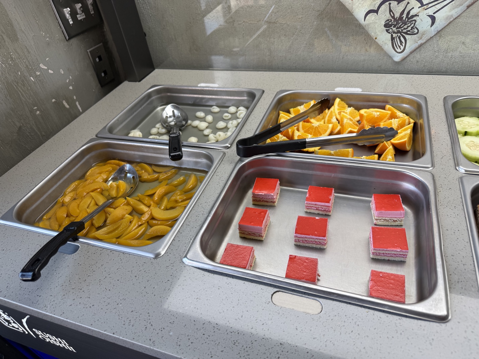 Fruit and dessert trays at the Sushi Oishii hot bar — sliced oranges, banana, and small layered cakes in steel pans