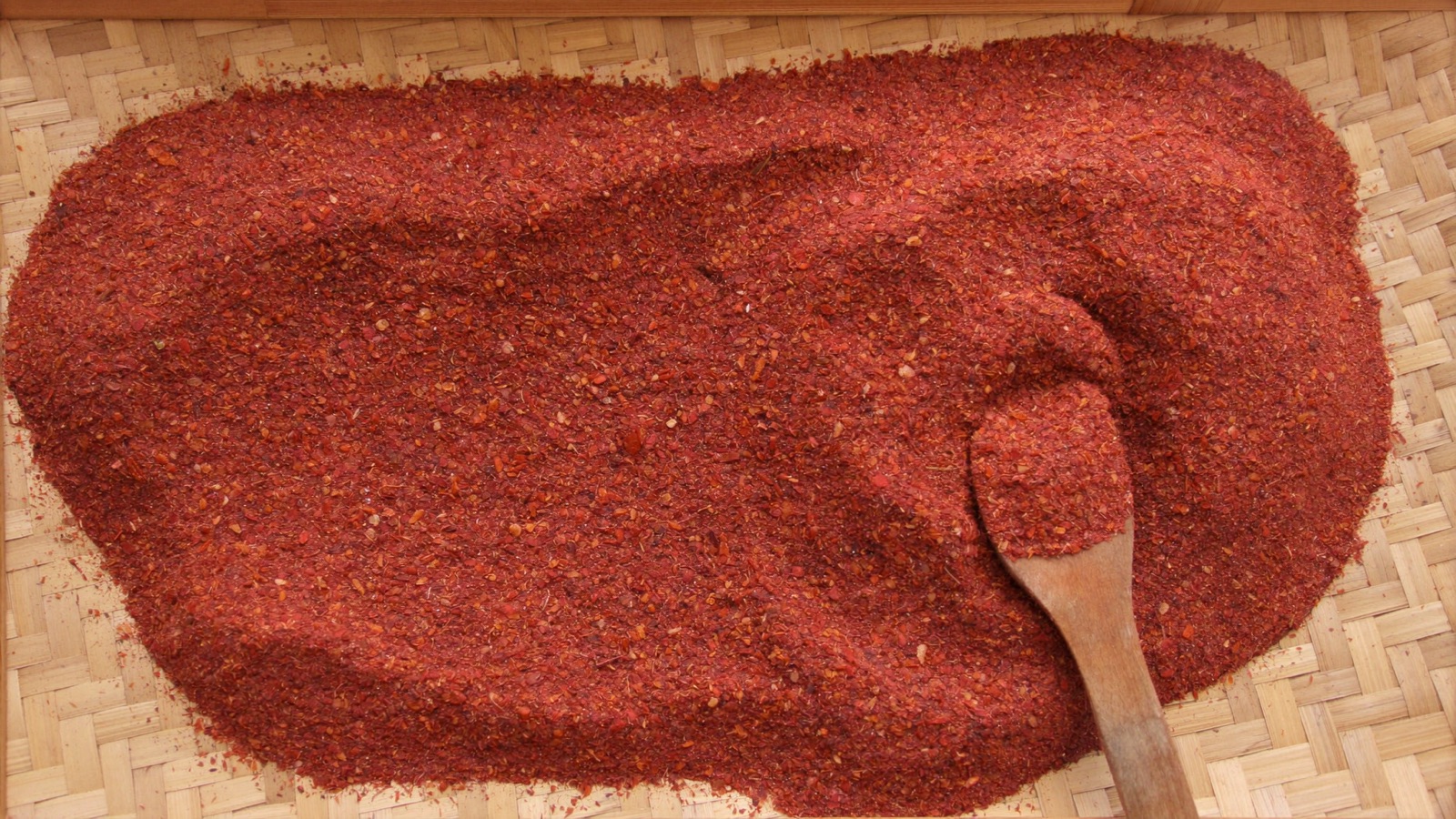 Coarse sun-dried Korean red chilli powder spread across a woven bamboo tray with a wooden scoop resting in it.