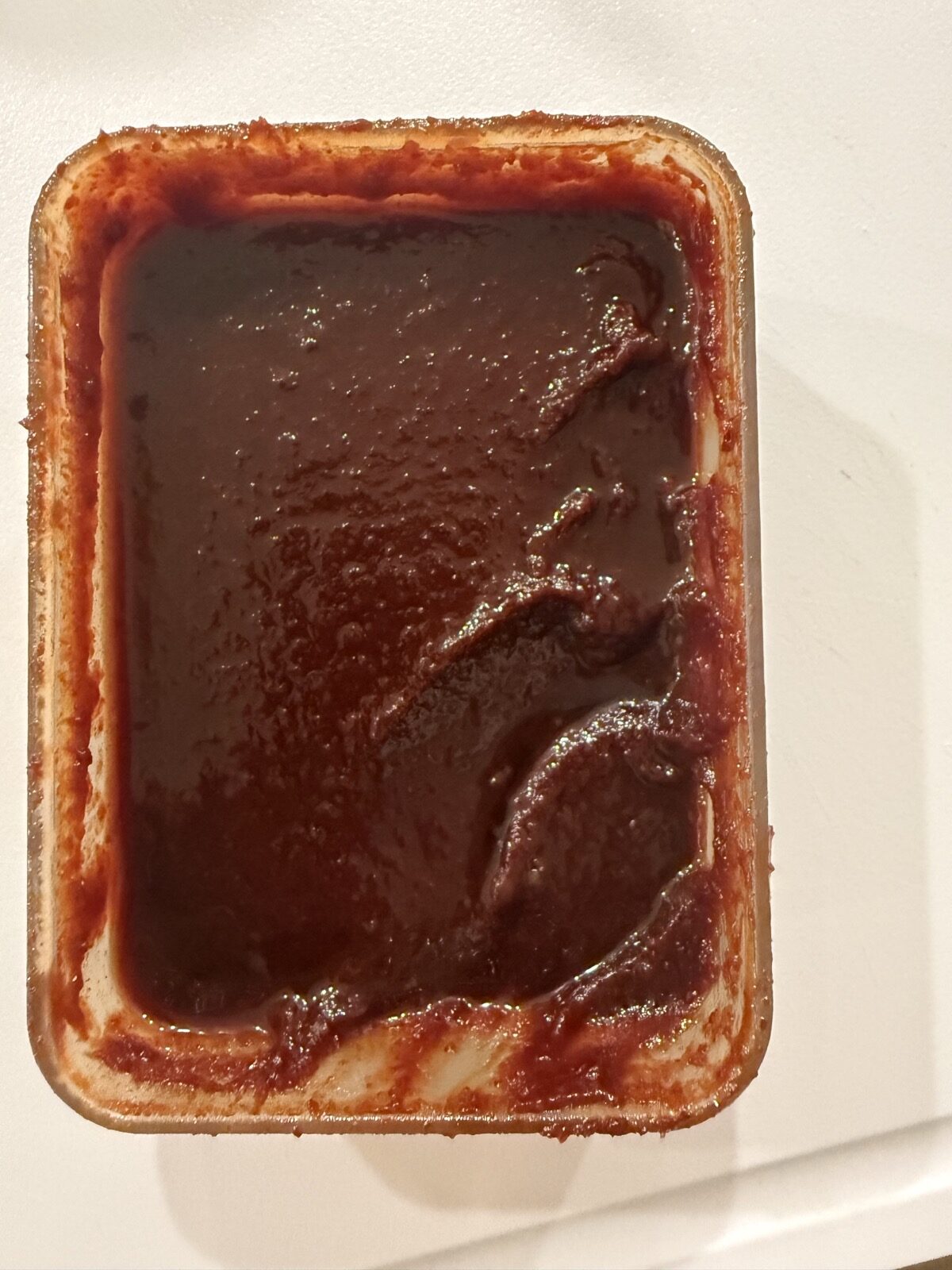 An opened tub of gochujang seen from directly above, its surface darkened after months in the refrigerator.